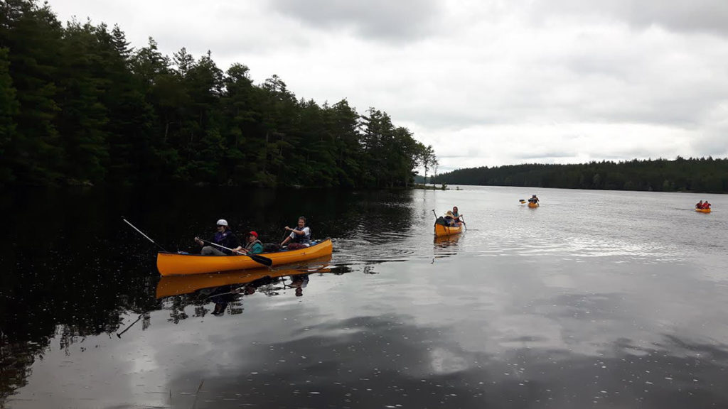 Out-tripping Adventure Camp - Orange canoes paddle in a row on gray water under a gray sky.
